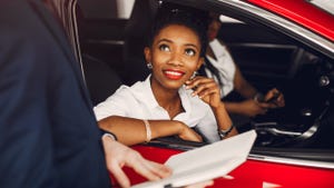 Two Black women sit in a red car at a dealership. There is a third person out of the frame holding documents.