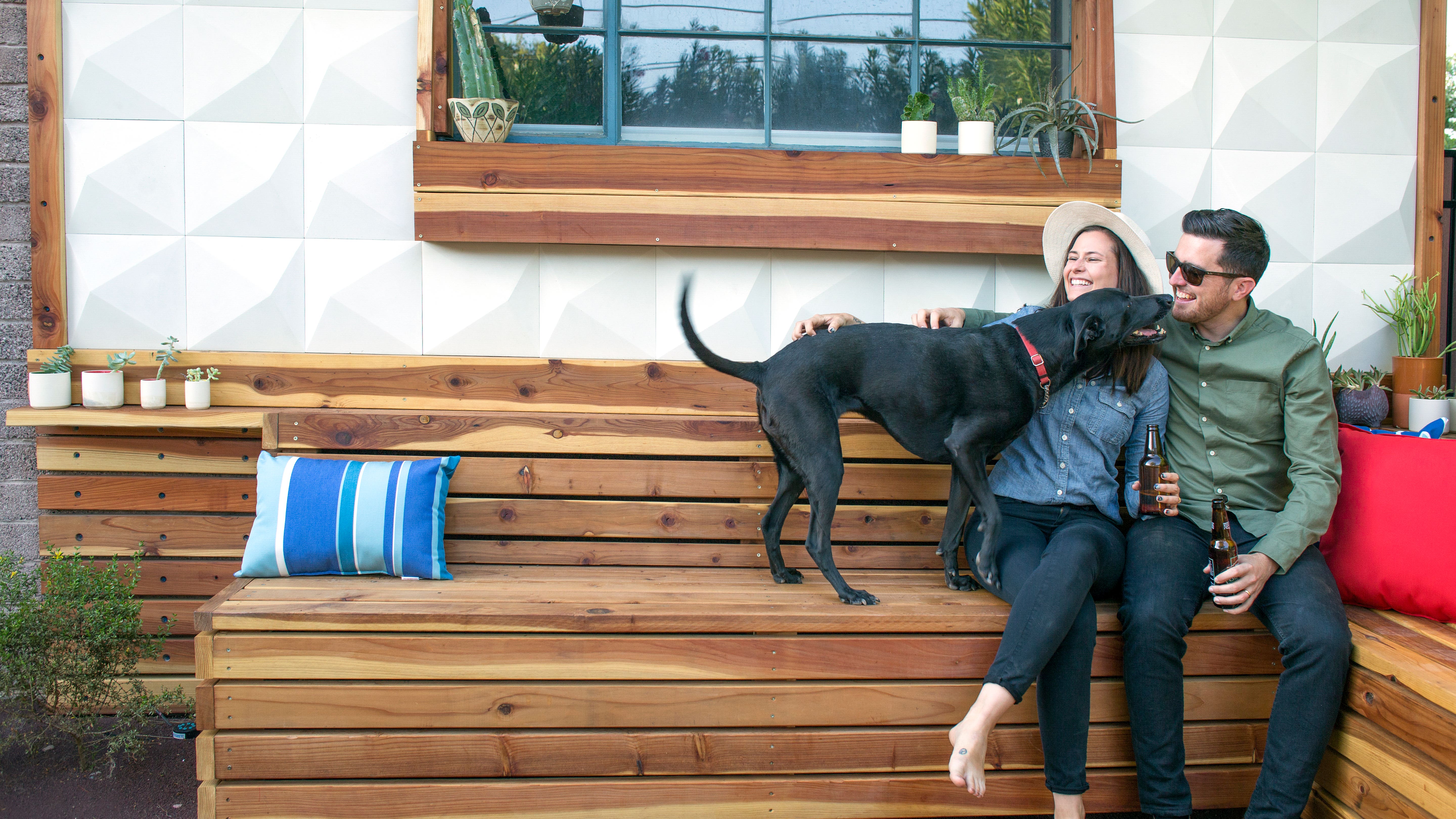 Happy couple holding beer bottles playing with dog while sitting in backyard of home