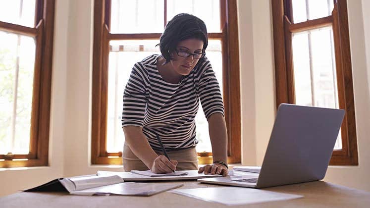 Woman leaning over and writing on paper | PeopleImages/DigitalVision/Getty Images