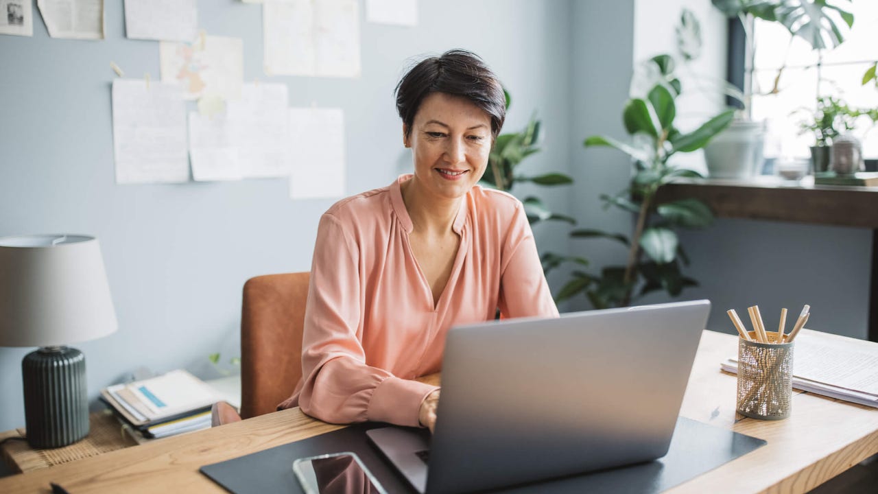 Middle aged woman smiles and uses laptop in home office at desk
