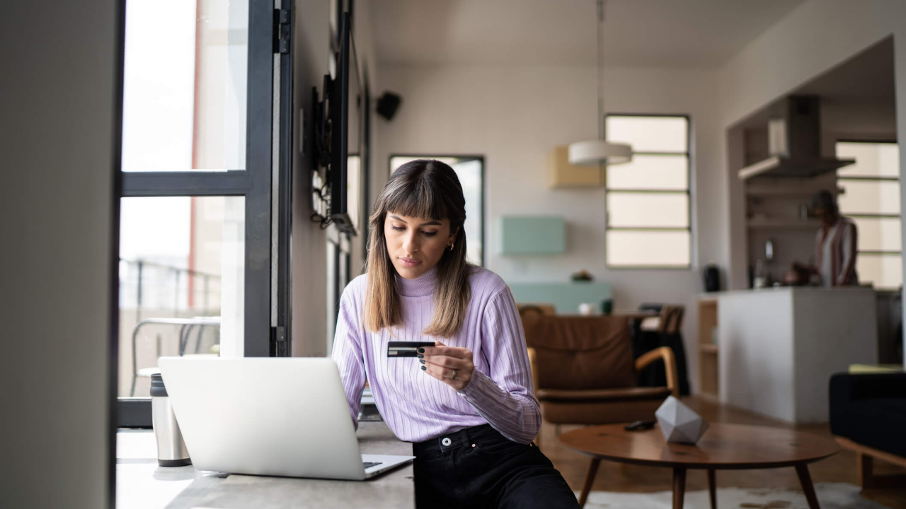 A young woman holds her credit card while working on a laptop in her apartment