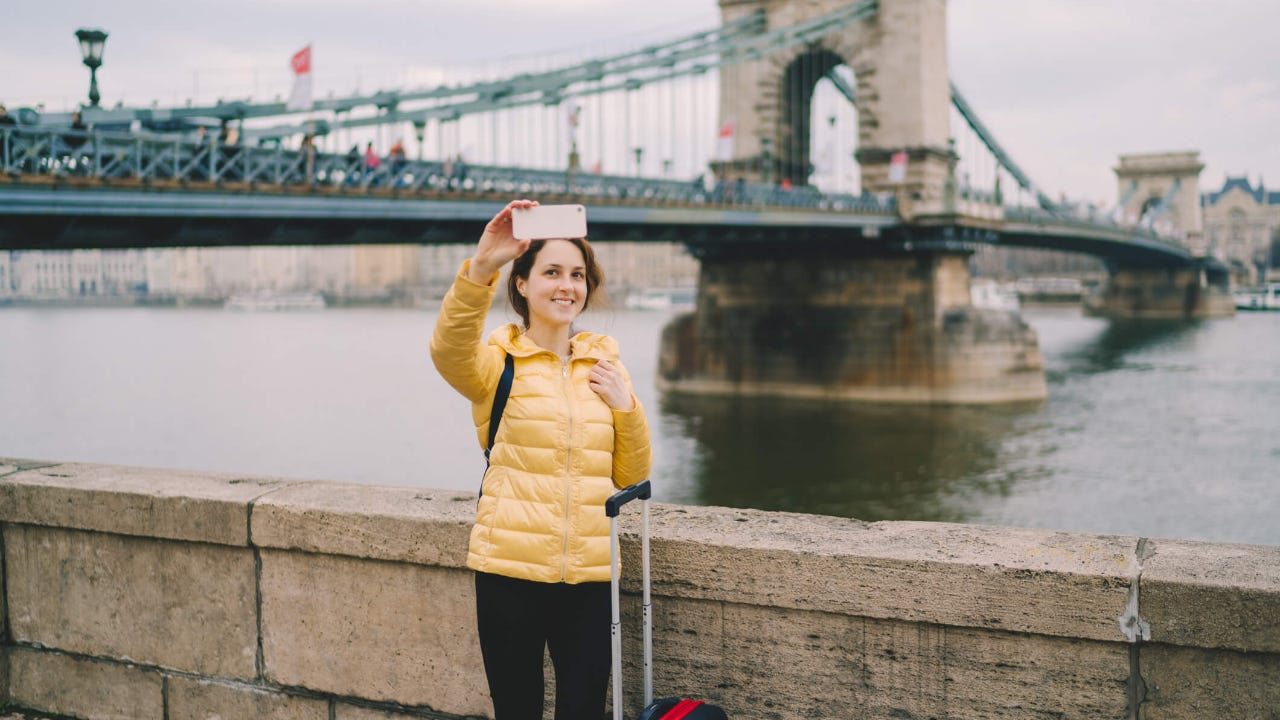 Tourist woman with suitcase takes selfie in front of a bridge in Budapest