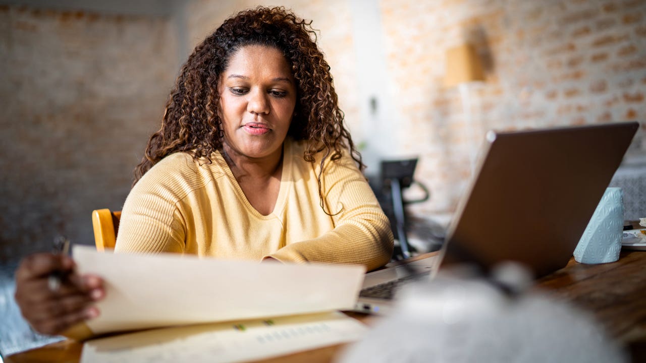 Black adult reviewing tax forms at their desk with an open laptop nearby.