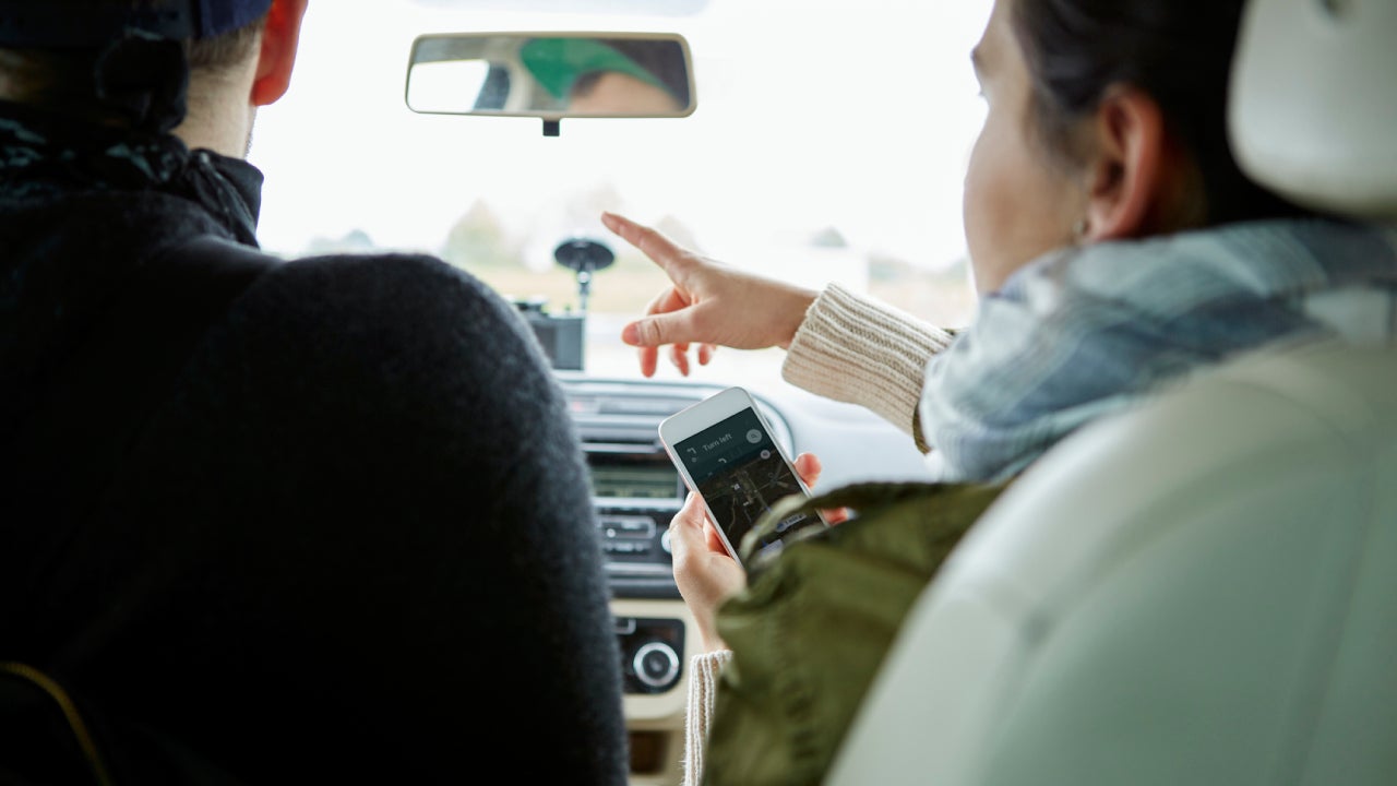 Young couple sitting in car, woman holding smartphone, pointing directions, rear view