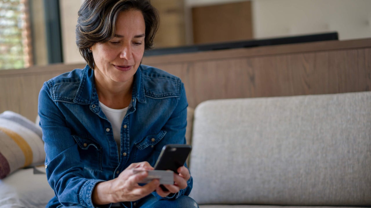Middle-aged woman uses her phone while holding her credit card at home