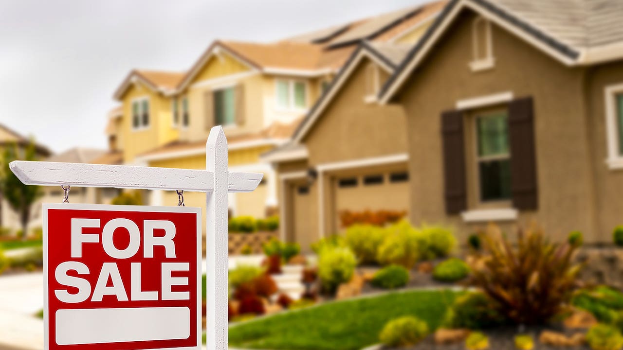 tan-colored suburban home with extensive landscaping and a red-and-white FOR SALE sign in front