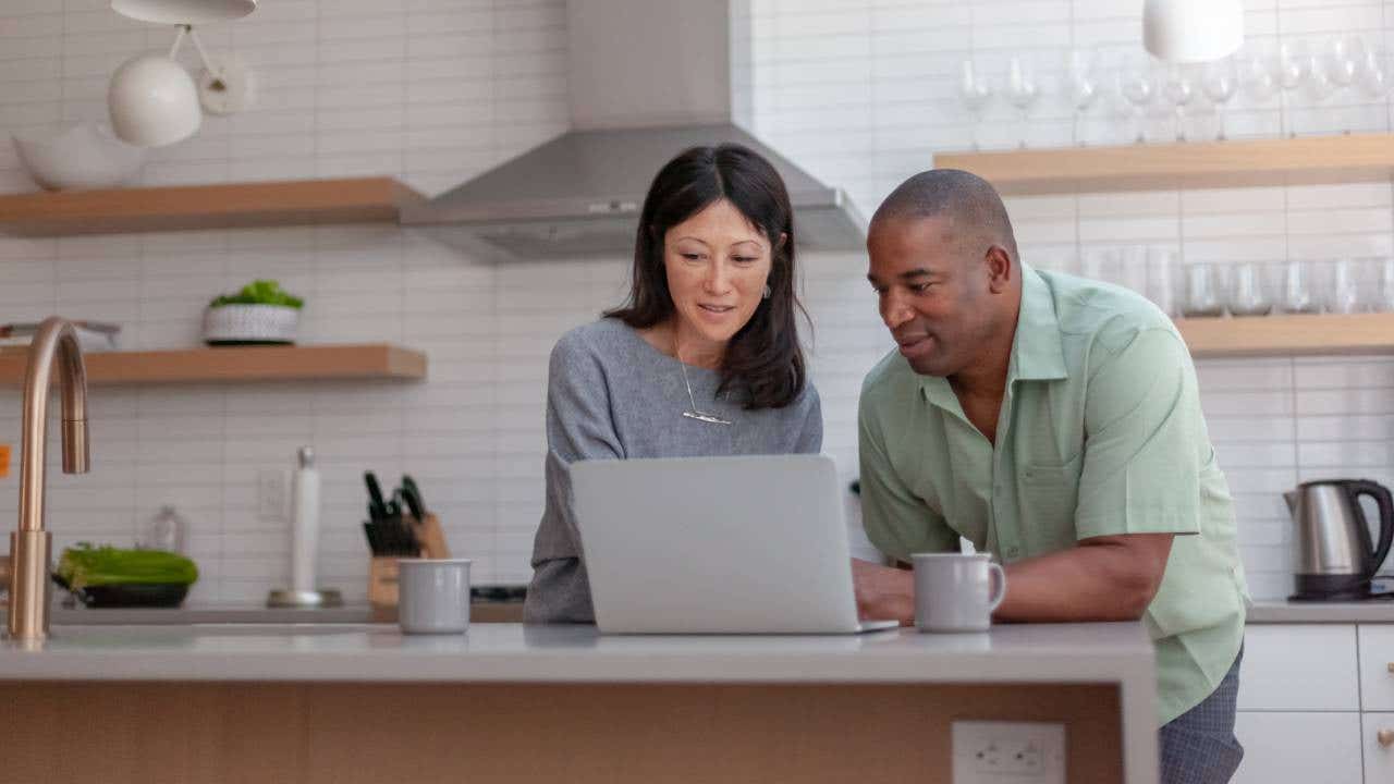 A mixed race couple use a laptop computer to pay bills online, manage budget and prepare tax documents. They are in the kitchen of their home.