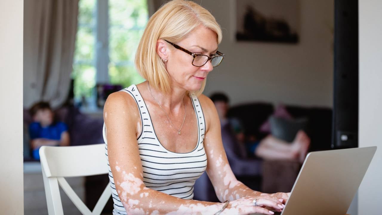 Blonde woman sitting her dining table at home with a laptop open in front of her as she works