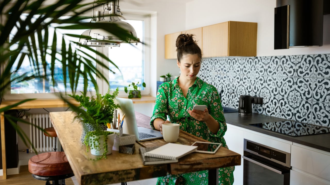While looking at her cellphone, a young woman sits at her kitchen table with a notebook, tablet, and laptop.