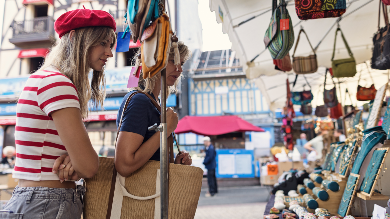 Two women are examining items for sale at an outdoor market in Etretat, Normandy, France. The young woman on the left is wearing a red beret, a red and white striped top. The mother, mature woman on the right, dressed in a navy-blue shirt and carrying a large straw bag.