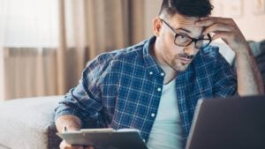 A confused man looks at his laptop while holding paperwork.