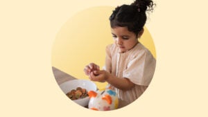 Image of a little girl holding coins in her hand. She sits in front of a table with a bowl full of coins and a multi-colored piggy bank.
