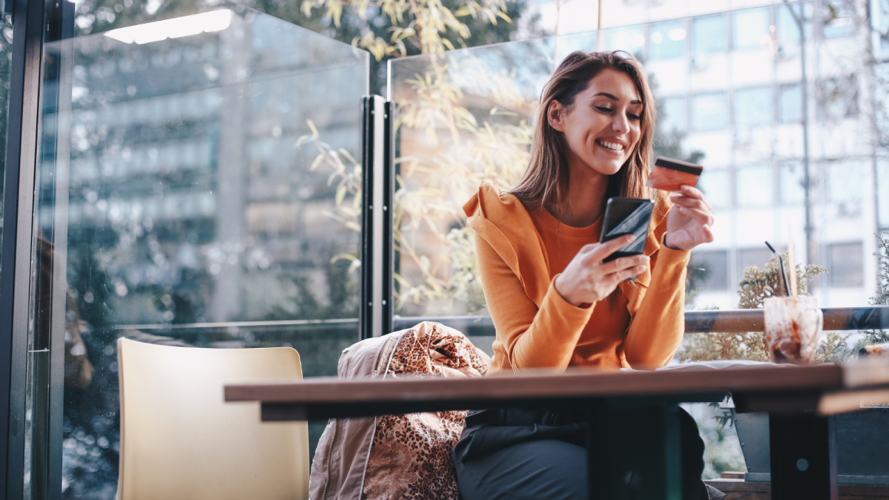 Young woman is buying online and paying with her credit card. She is sitting at the café, and enjoying her day alone. Because it is COVID-19 epidemic, she decided to sit alone. On table is protective face mask and knit hat she wore when she walked out of house.