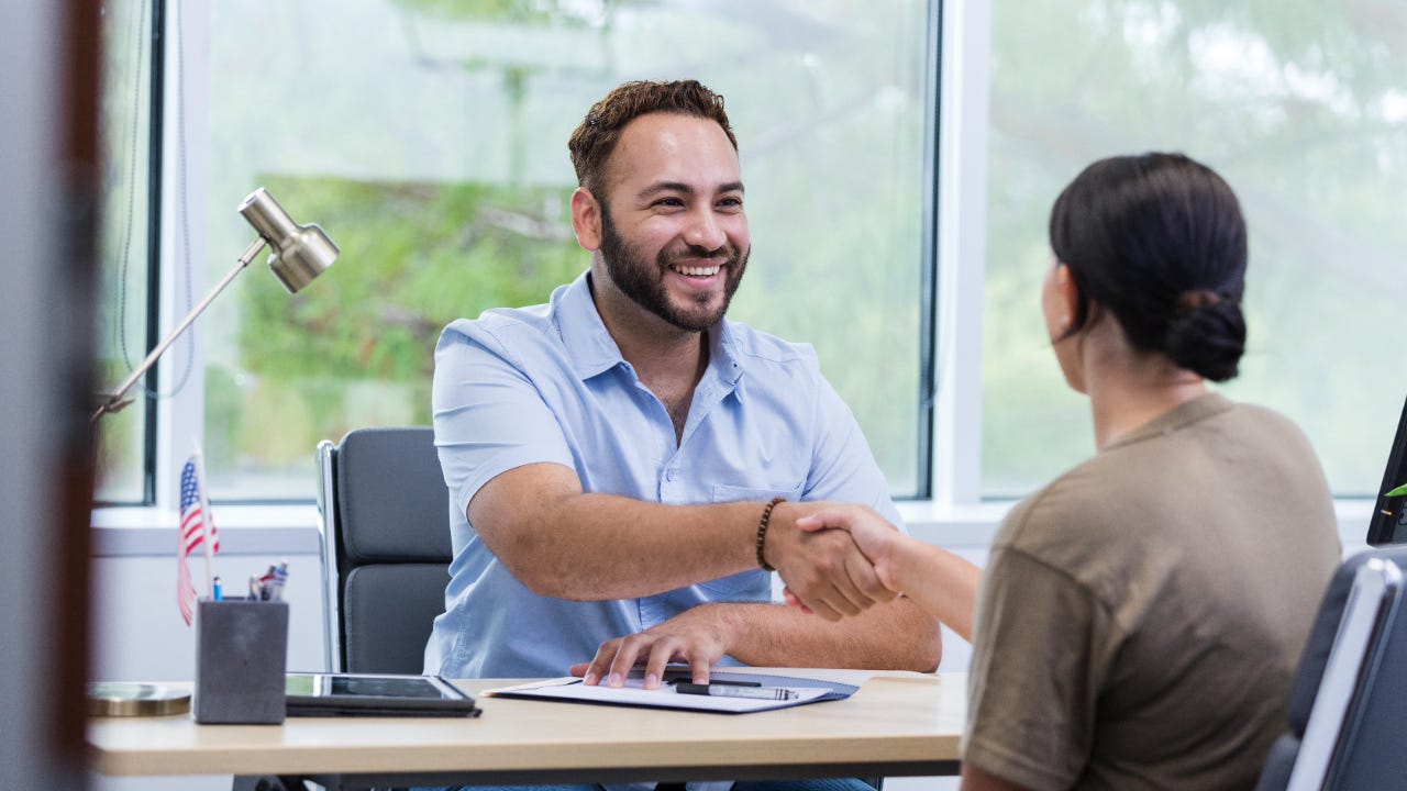 Smiling male shakes hands over his desk with a woman in a brown shirt who is facing away from the camera. They are in a credit union office.