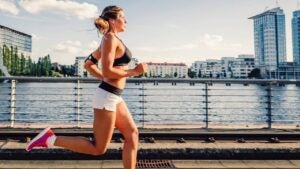 A picture of a women running and exercising beneath a clear sky, along the spree river in berlin. She is wearing sport dress and listen to music over headphones. In the distance below are some buildings and the sun is setting giving a nice warm light.