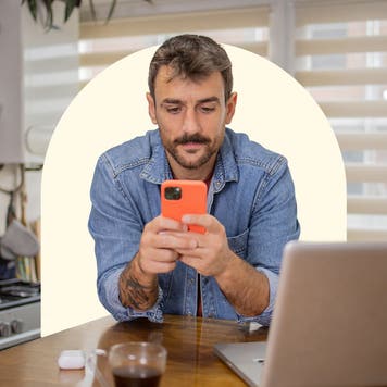 A white man sits at a kitchen table while using an orange phone. There is a laptop open in front of him. Design includes a light yellow semicircle behind him.