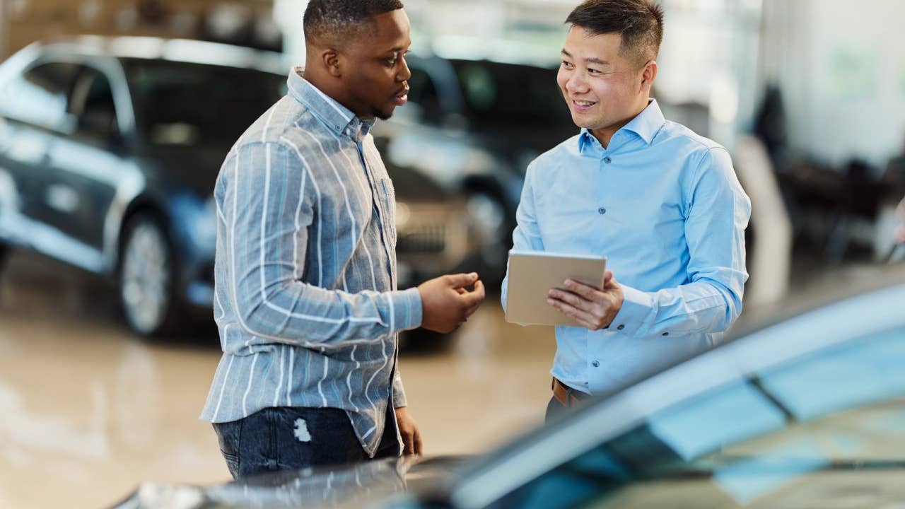 Smiling Asian salesman talking to a Black customer while using digital tablet in a car showroom.