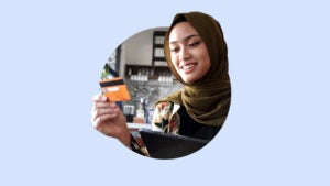 Smiling Muslim woman in a hijab looks at her credit card. Image is on top of a light blue-purple background.