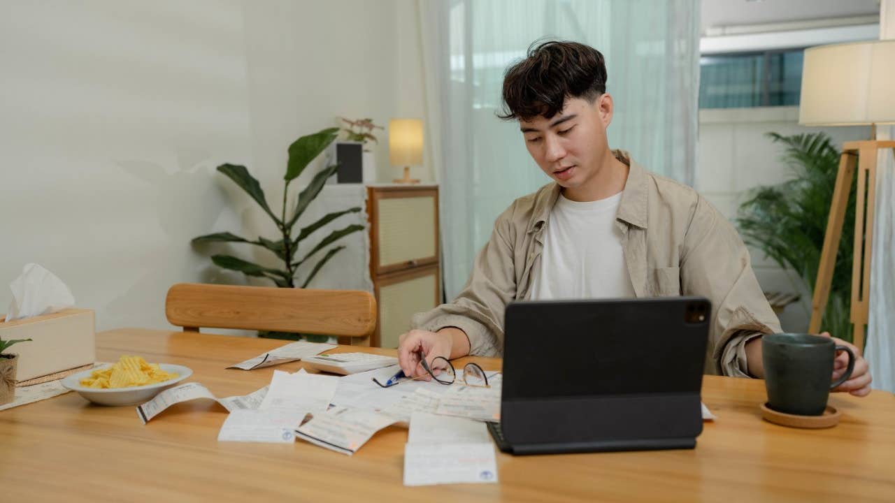 Asian male sits at dining room table with laptop and financial reports doing monthly budget to do monthly finances, pay taxes and save money for the future.
