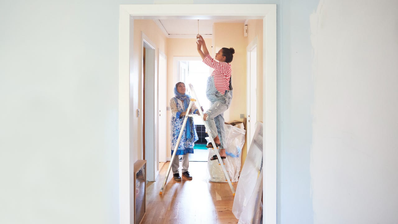 Older woman holds a ladder for a younger woman while she changes a light fixture.