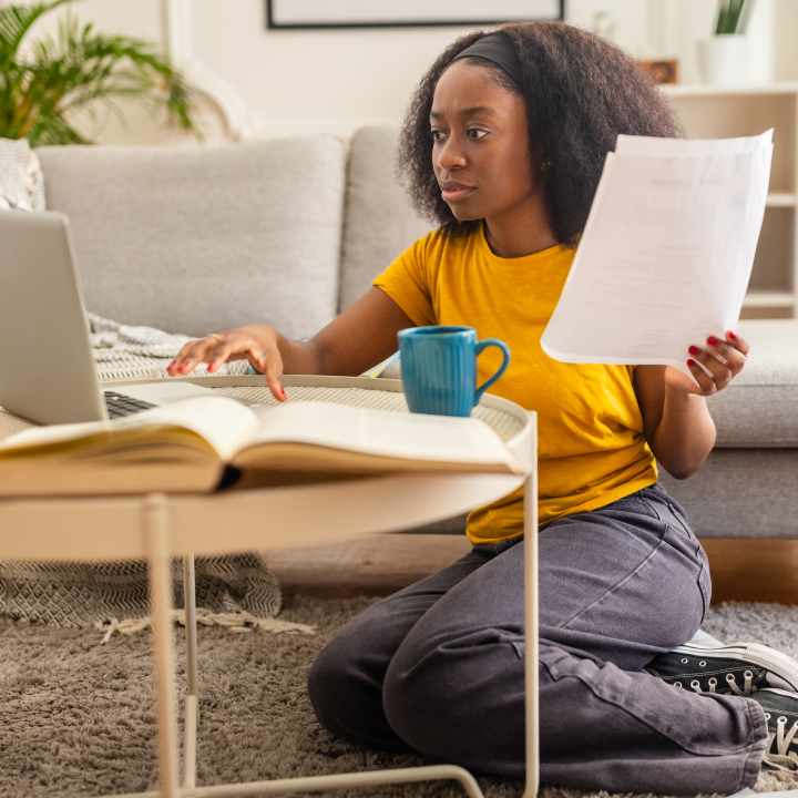 A focused woman in a yellow shirt examines a bill while working on her laptop, sitting on the floor with various documents around her, in a welcoming living room setup