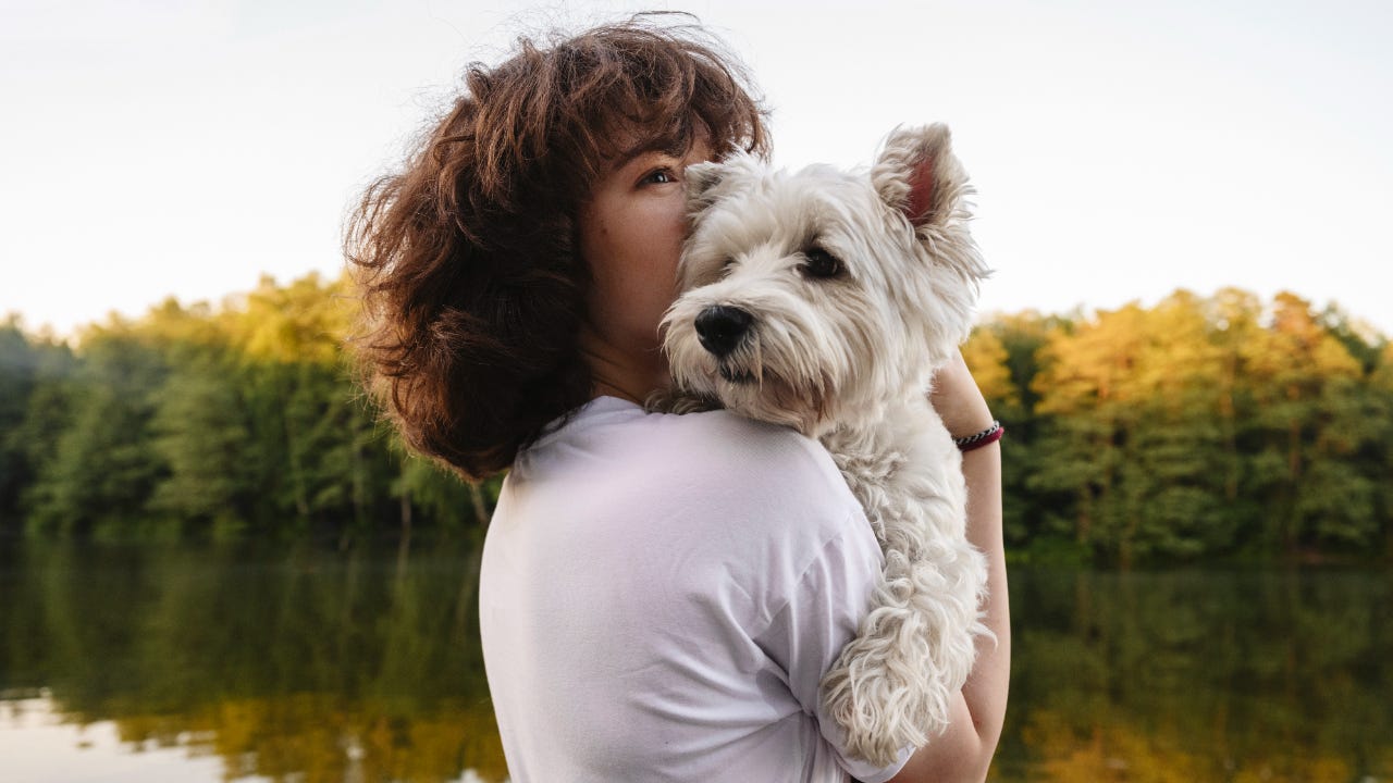 A woman stands beside a lake holding a white puppy.