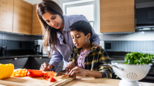 Mother helping young son cut vegetables in kitchen.