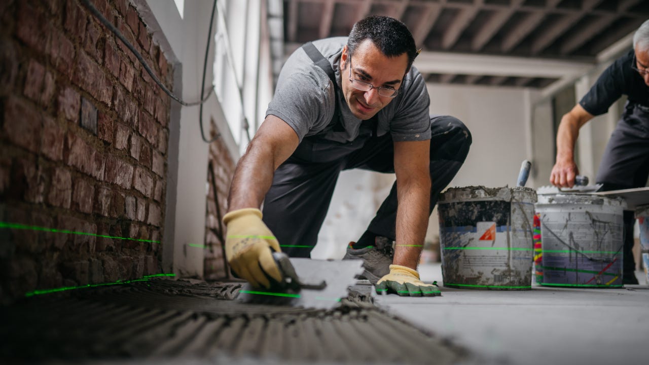 A man kneels on the floor and spreads mortar on the floor in preparation for laying tile.