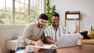 Young multiracial gay couple smiling while going over their home finances together at a table in their living room at home.