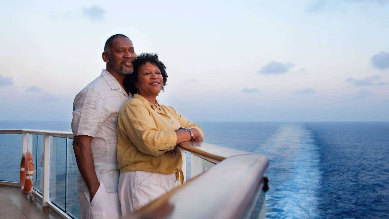 Mature African American couple, standing and leaning on railing on deck of a cruise ship, looking off into the distance.