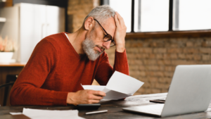 Sad, depressed caucasian man holding documents, sitting at computer in home office.