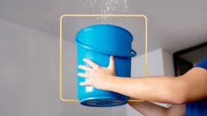 Someone offscreen holds a blue bucket up to a leaking ceiling. There is an illustrated yellow square in the background.
