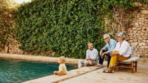 Wide shot of smiling grandparents relaxing with son and granddaughter beside pool at tropical villa during multigenerational family vacation