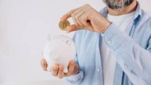 A man with a gray beard holds a bitcoin token over a piggy bank.