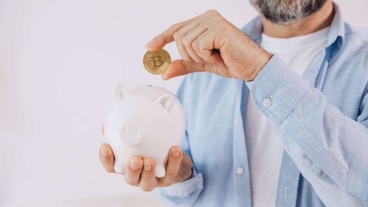 A man with a gray beard holds a bitcoin token over a piggy bank.