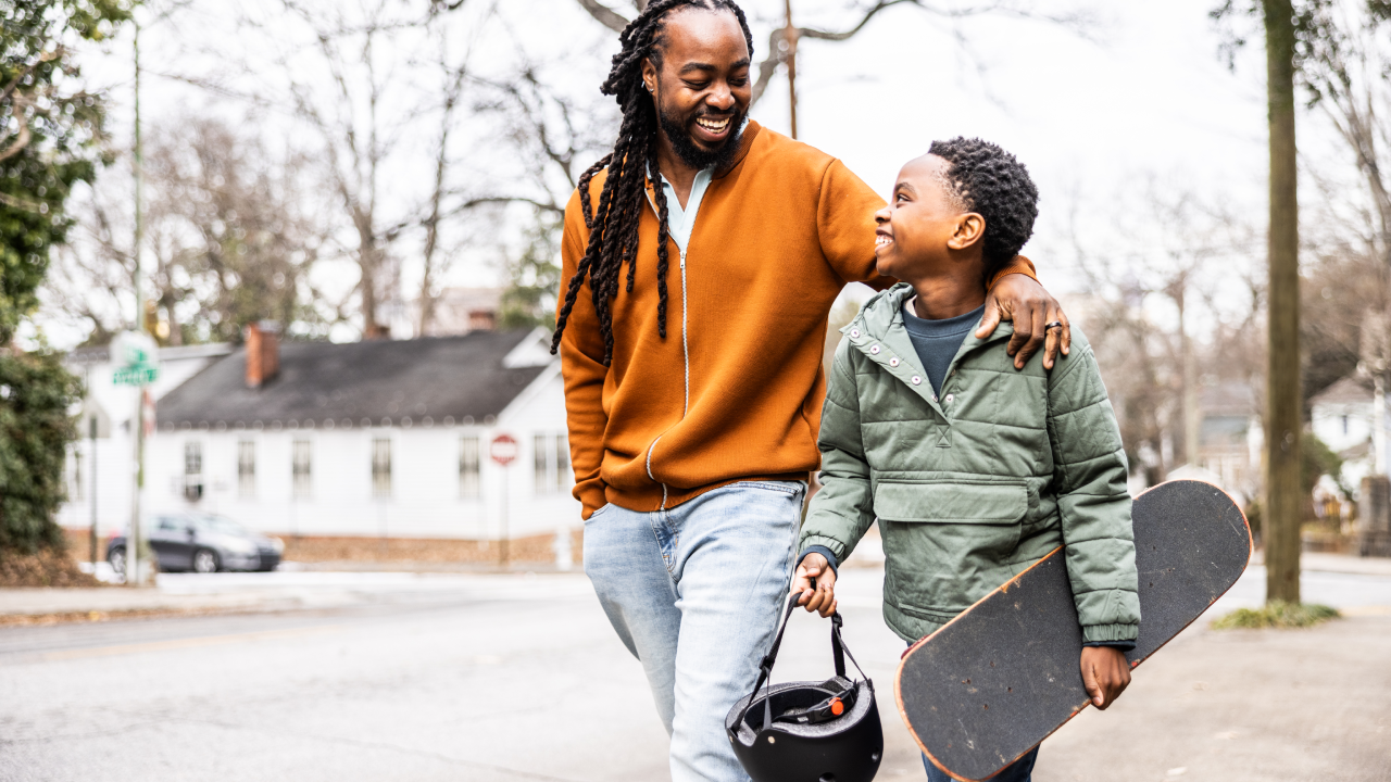 Father and young son with skateboard walking down the street.