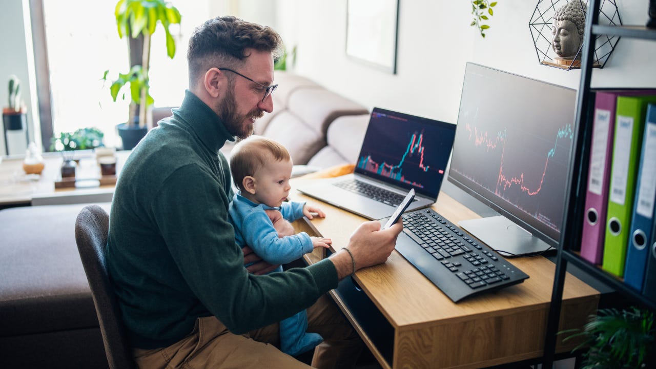 Man looking at currency trading app on his smart phone from his home office while holding a baby on his lap.