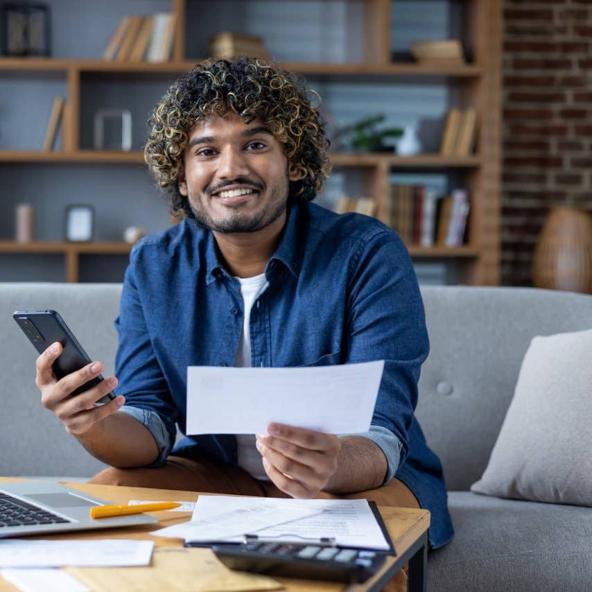 A smiling man reviews financial documents while holding a phone, seated on a couch in a cozy living room setting.