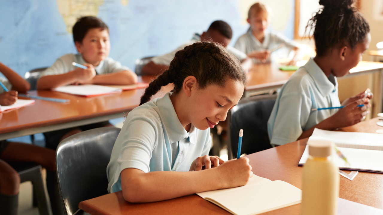 Girl with braided hair writing in book while sitting at desk with students in classroom.