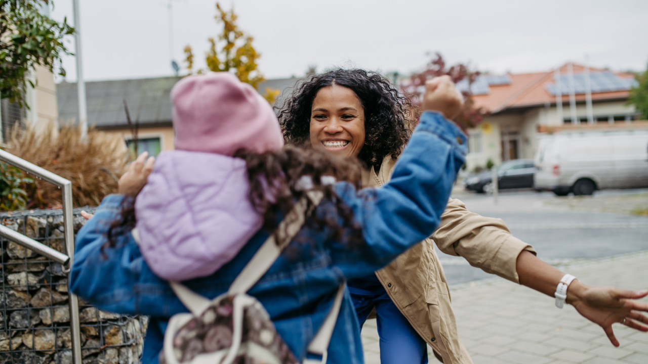 Mother picking up small daughter from school, embracing in front school building.