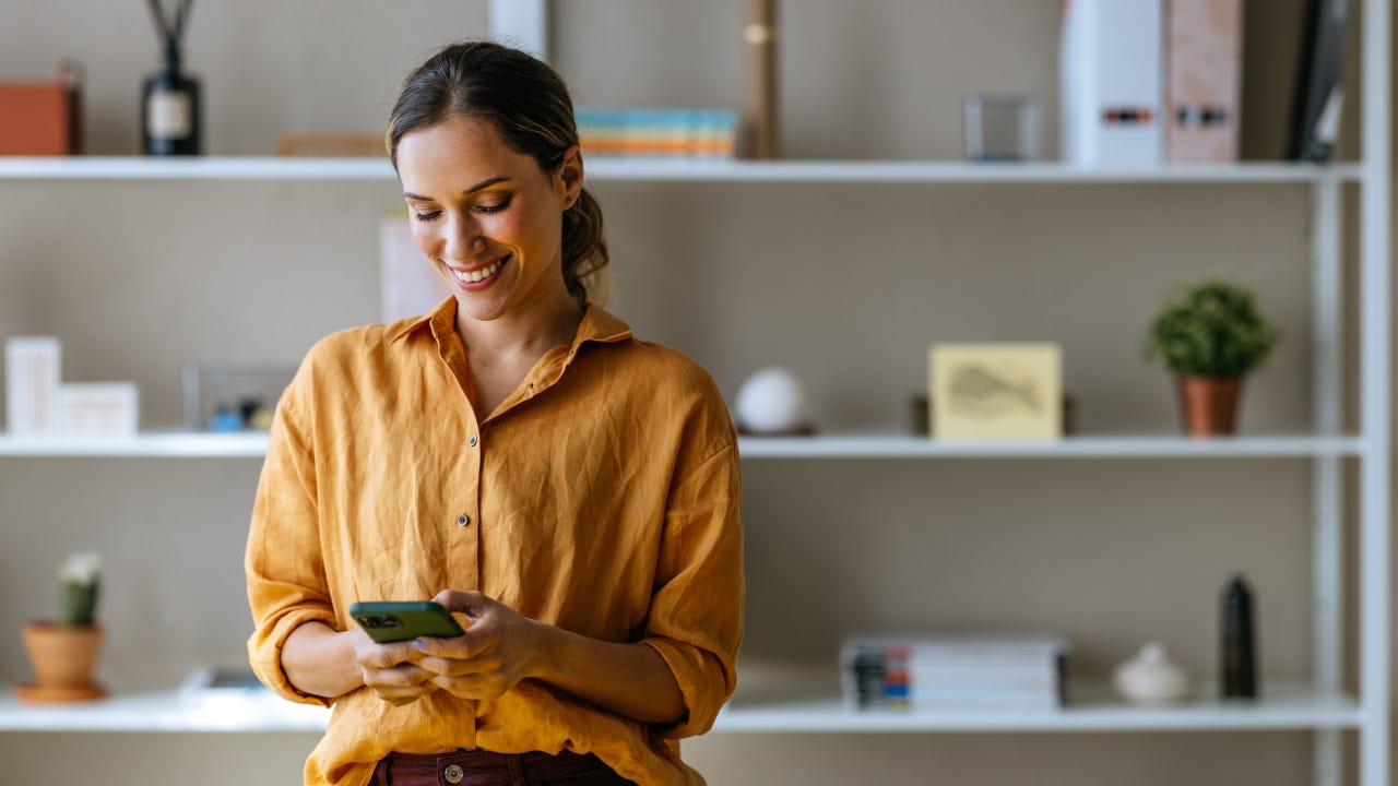 A woman looks at her investing portfolio on her smartphone in a living room with shelves behind her